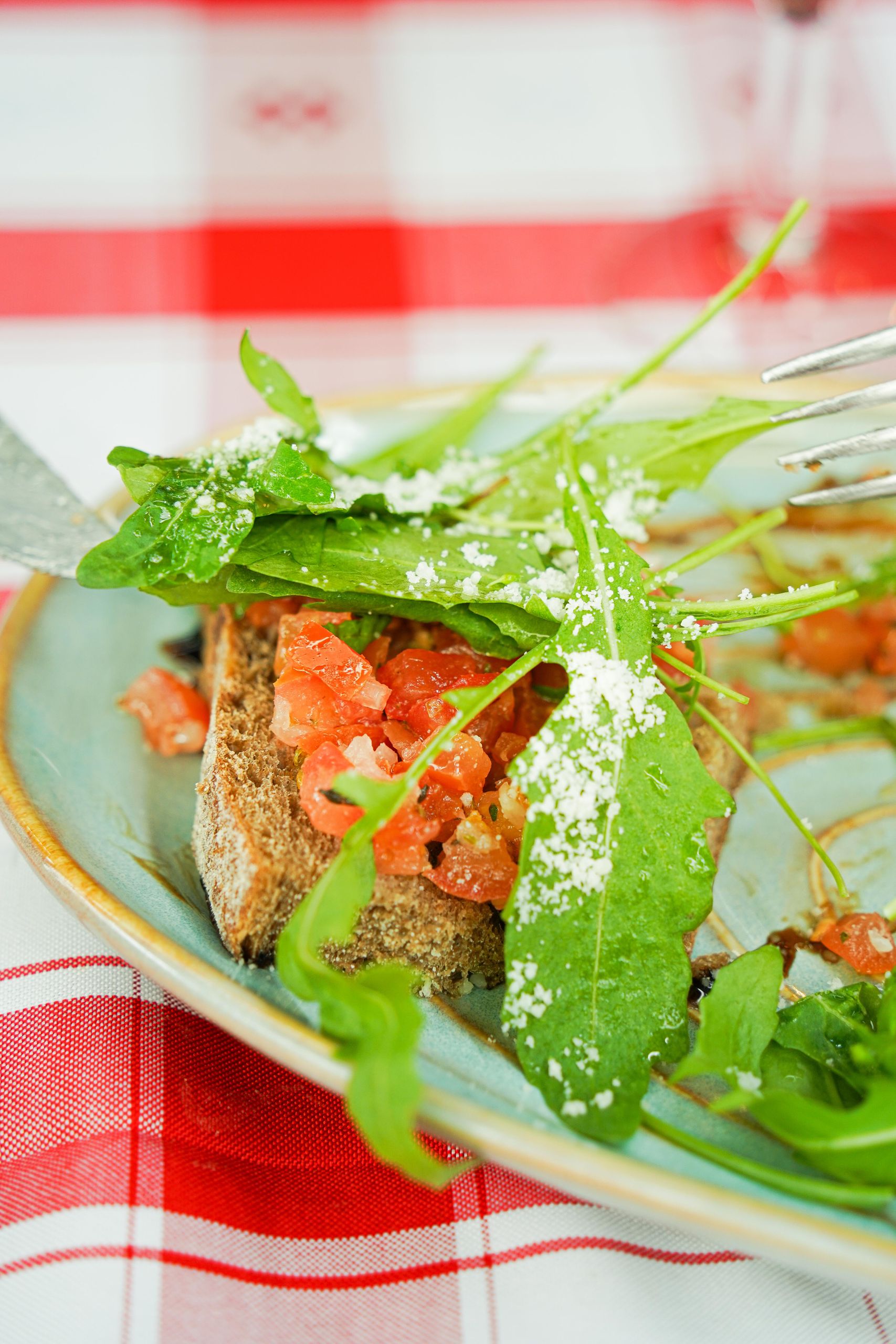 Close-up of bruschetta topped with arugula and parmesan on a checkered tablecloth.